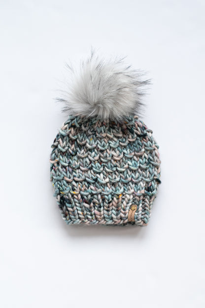 Blue hand knitted beanie made with chunky yarn with large fluffy gray faux fur pom pom on a white background