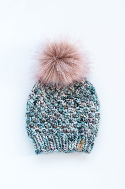 A hand-knit merino wool hat in blue speckle pattern with a faux fur pom pom on top.