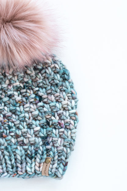 Close up view of a blue textured knit beanie with a fluffy pink faux fur pom pom