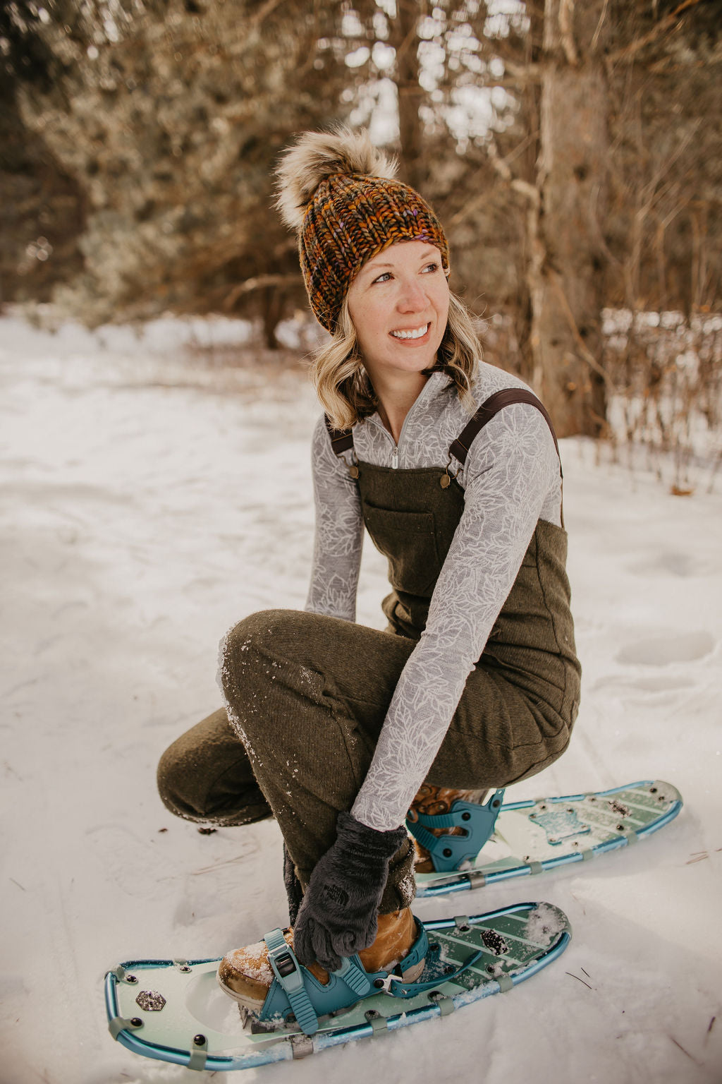 Woman wearing brown ribbed knit hat and green overalls bends to adjust snowshoes in a snowy forest