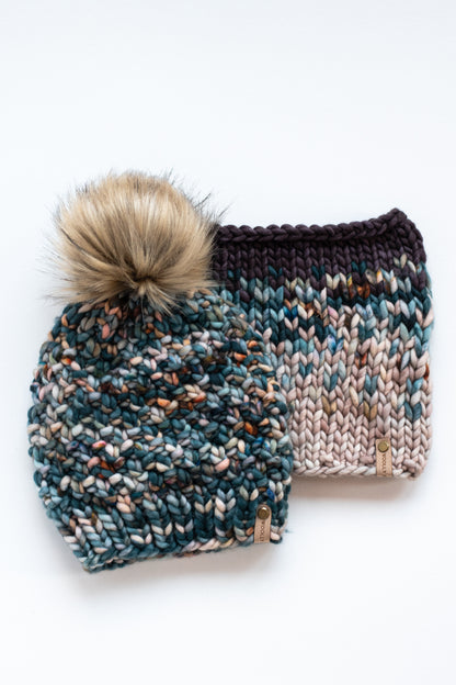 A multicolor green hand knit hat with fluffy pom pom and colorful knit cowl on a white background.