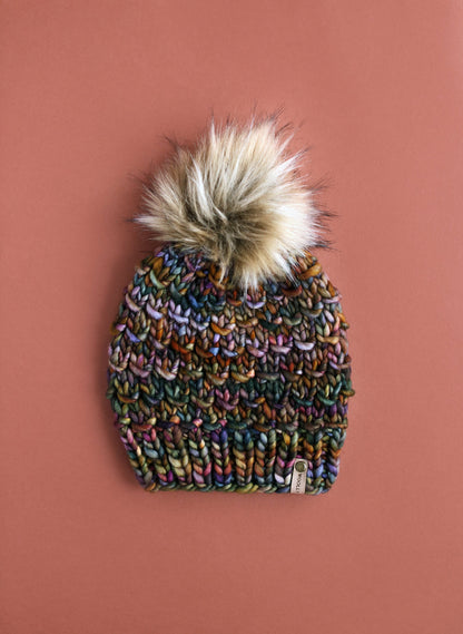 Brown multicolor knit beanie with fluffy brown faux fur pom pom on a brown background. 