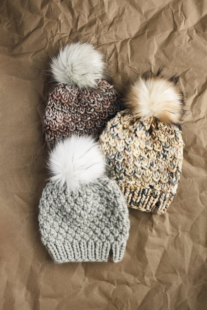 Stack of three neutral colored knit hats lays on a brown kraft paper background