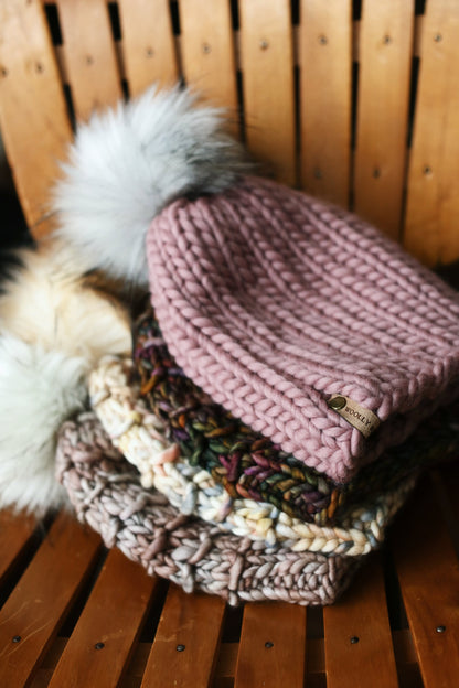 A stack of chunky knit beanies with pom poms sits on a wooden chair