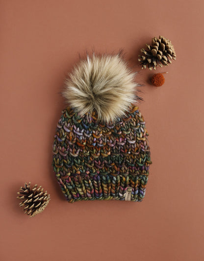 Brown colorful chunky knit hat with fluffy brown fur pom pom on a brown background with pinecones.