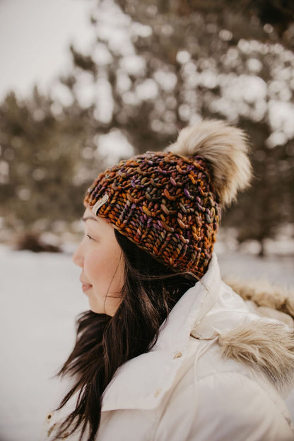 Woman with long dark hair wears a white jacket and a brown multicolor knit hat with fluffy pom pom in a snowy forest setting