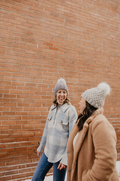Two women laugh while wearing coats and knitted pom pom hats in a brick alley
