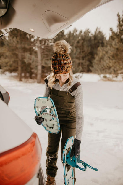 Woman wearing brown merino wool beanie and green wool overalls puts snowshoes into car in a snowy setting