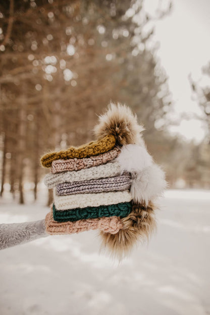 A person holds a stack of knitted hats with pom poms in a snowy setting