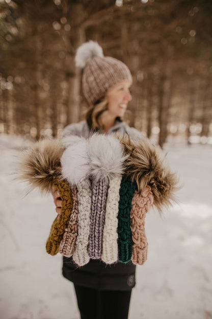 Woman looking away from the camera holds a stack of seven knit hats in a snowy forest