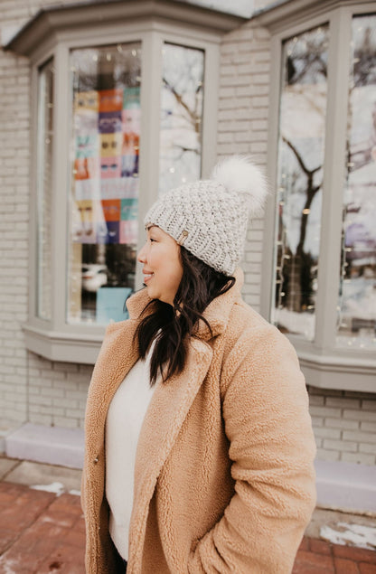 Woman wears a fuzzy brown coat and light gray hat standing in front a shop window