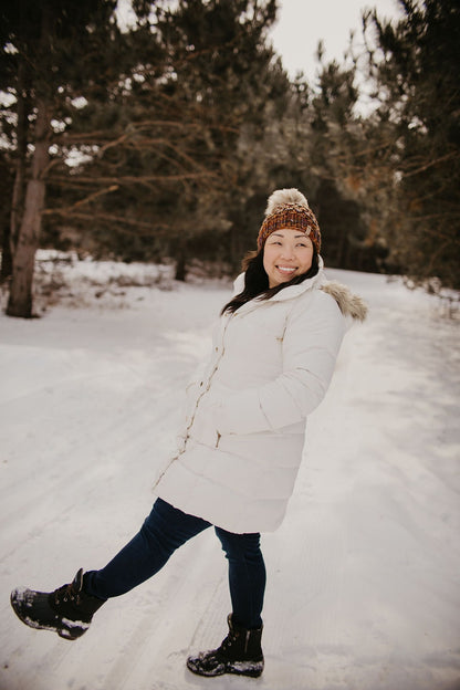 Woman with long dark hair wears a white jacket and a brown multicolor knit hat with fluffy pom pom in a snowy forest setting