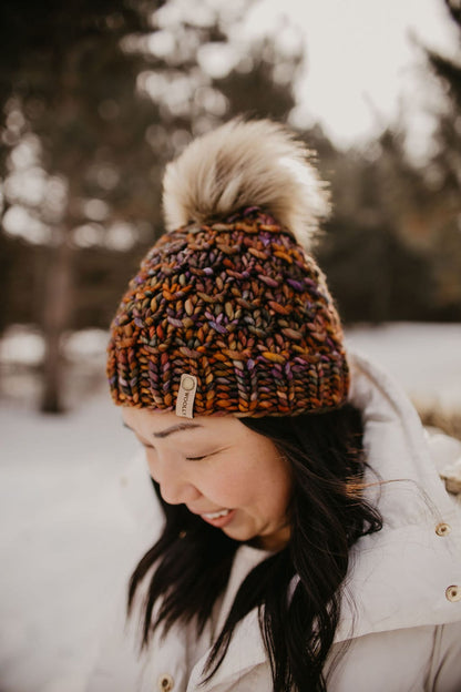 Woman with long dark hair wears a white jacket and a brown multicolor knit hat with fluffy pom pom in a snowy forest setting