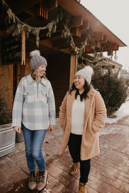 Two women wear hand knit beanies in front of a store decorated with pines in the winter