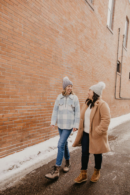 Two women wearing coats and knitted pom pom hats walk down alley with brick background