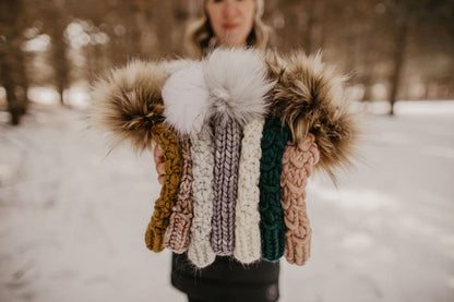 Woman holds stack of six chunky knitted hats with pom poms in a snowy forest