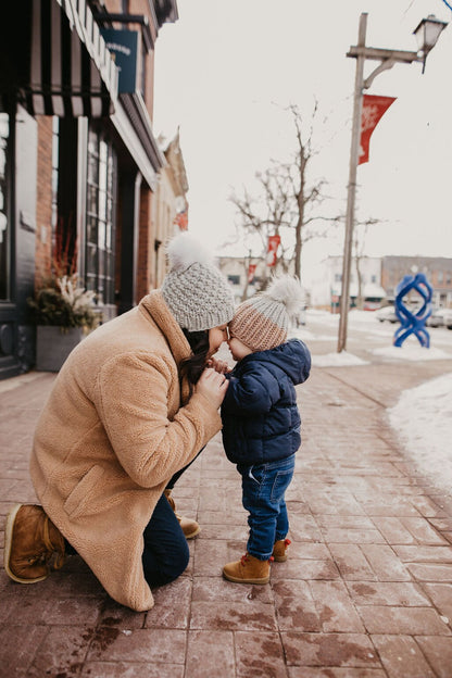Woman wearing brown fuzzy coat and knit hat kneels down to hold hands with a toddler wearing a knit hat