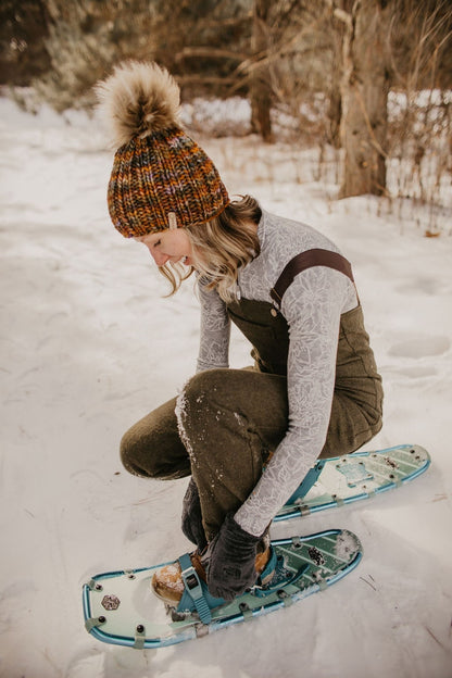 Woman wearing brown ribbed knit hat and green overalls bends to adjust snowshoes in a snowy forest