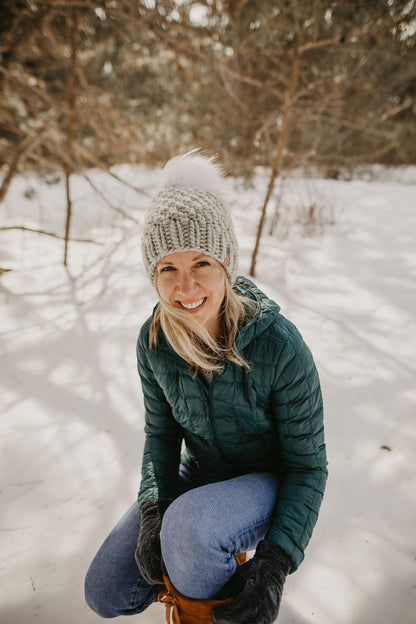 Woman kneels in the snow to adjust her boot wearing a green puffer jacket and light gray knitted hat