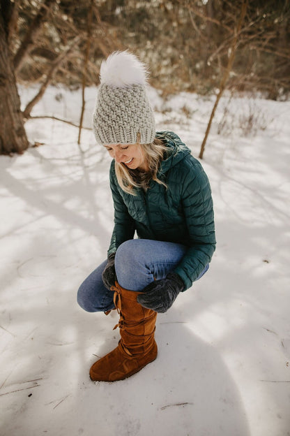 Woman kneels in the snow to adjust her boot wearing a green puffer jacket and light gray knitted hat