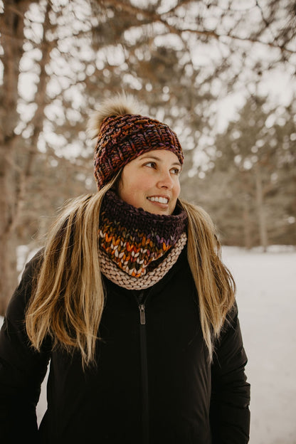Woman wears a black jacket with brown colorful knit hat and cowl in a snowy forest setting