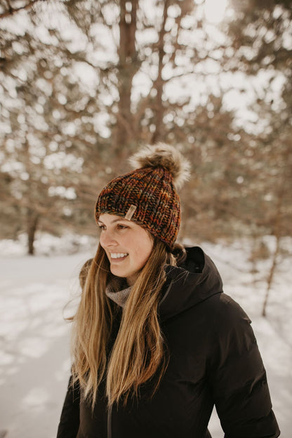 Woman wears a black jacket and brown chunky knit beanie in a snowy forest setting