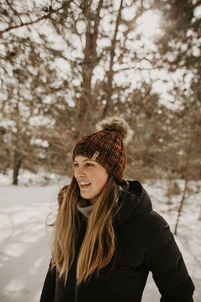 Woman with long brown hair wears a black jacket and brown colorful knitted beanie in a snowy forest.
