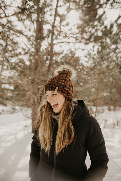 Woman wearing a brown ribbed knitted beanie and black jacket looks to the side laughing in a snowy forest setting