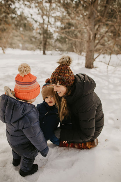 Woman kneels in snow wearing winter clothing to play with two young children in snowsuits.